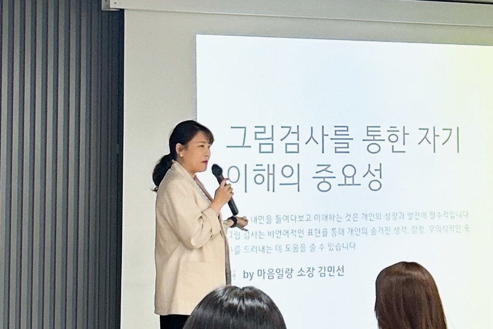 ‘그림검사를 통한 자기 이해의 중요성’이라는 화면 앞에서 마이크를 들고 강의하는 김민선 소장과 듣는 사람들의 뒷모습